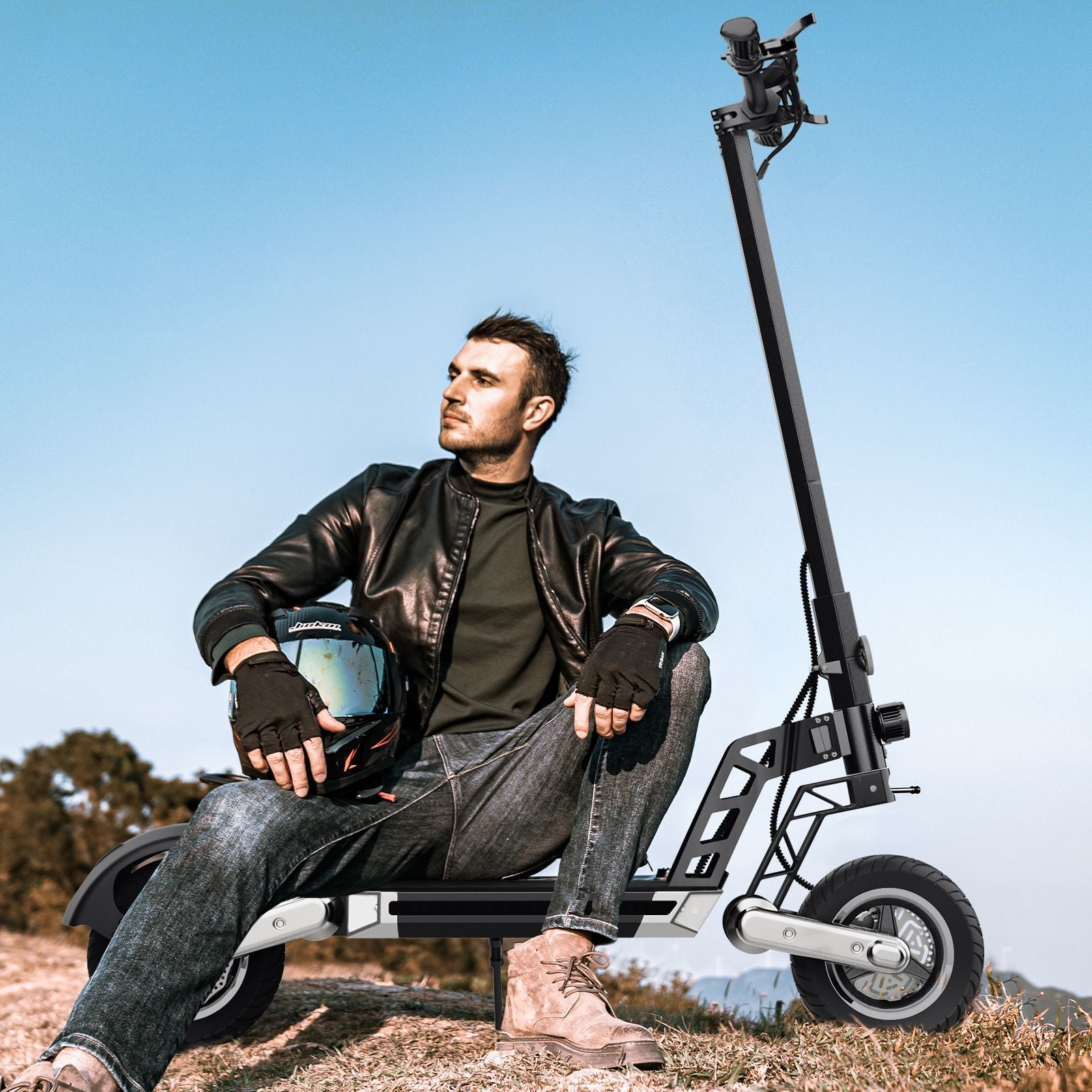 On a hill, against a backdrop of withered yellow meadows, a man in hiking boots sits on a silver-black Todimart-G2 ABE electric scooter (front and rear disc brakes with off-road tires), holding a helmet. Wearing a leather jacket, he exudes a carefree and relaxed vibe in the mountains.